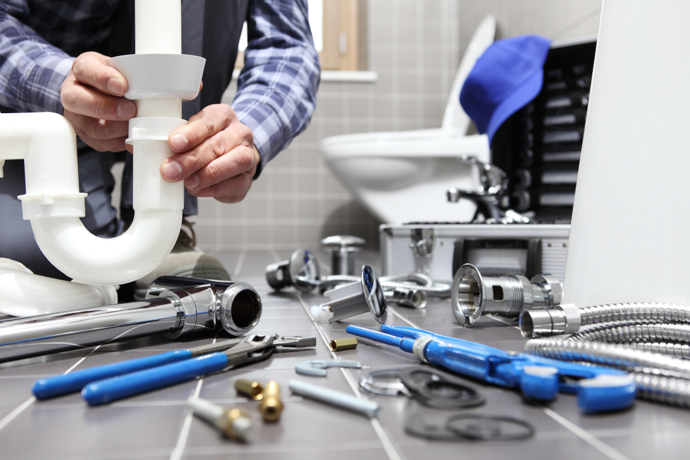 A person working on plumbing with tools and parts spread out on a bathroom floor.